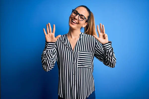 Beautiful blonde woman with blue eyes wearing striped shirt and glasses over blue background showing and pointing up with fingers number nine while smiling confident and happy.
