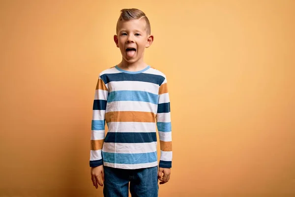 青い目の若い白人の子供は 黄色の背景にカラフルな縞模様のシャツを着ていて 面白い表情で幸せに舌を突き出しています 感情概念 — ストック写真