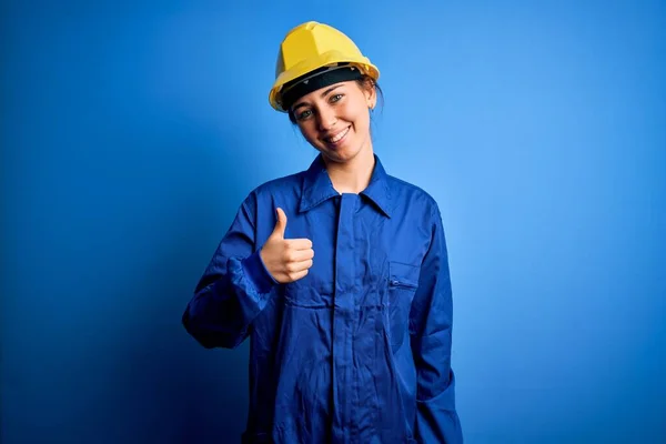 青い目の若い美しい労働者の女性は セキュリティヘルメットと手で幸せな親指アップジェスチャーを行う制服を身に着けています 成功を示すカメラを見て表現を承認する — ストック写真