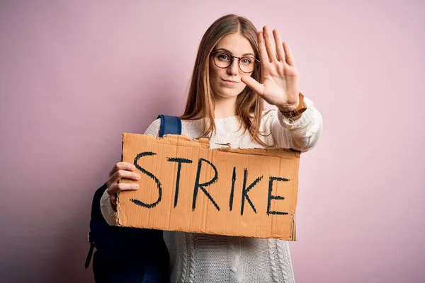 Young beautiful redhead activist woman holding banner with strike message with open hand doing stop sign with serious and confident expression, defense gesture