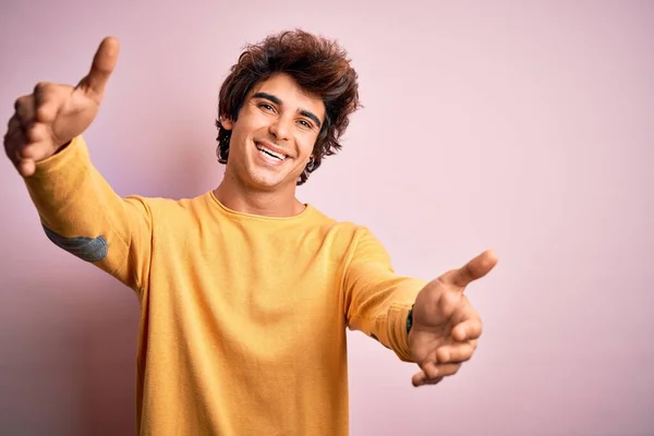 孤立したピンクの背景の上に立って黄色のカジュアルなTシャツを着て若いハンサムな男は抱擁のためのオープン腕で笑顔カメラを見て 幸せを受け入れる陽気な表情 — ストック写真