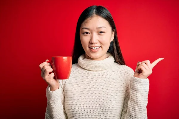 年轻的亚洲女人在孤立的红色背景下喝着一杯热咖啡 她非常高兴地将手和手指指向旁边 — 图库照片