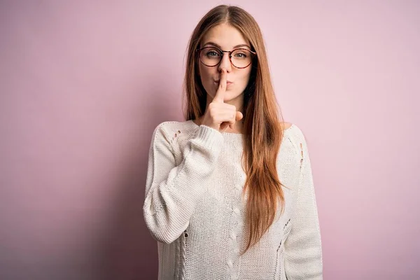 ピンクの背景にカジュアルなセーターと眼鏡を着た若い美しい赤毛の女性は 唇に指で静かになるように求めています 沈黙と秘密の概念 — ストック写真
