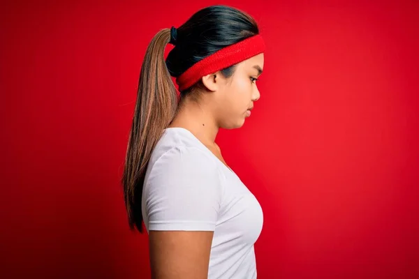 年轻美丽的亚洲女运动员身穿运动服 身披红色背景的运动服 面向侧面 带着自信的微笑放松自己的姿态 — 图库照片