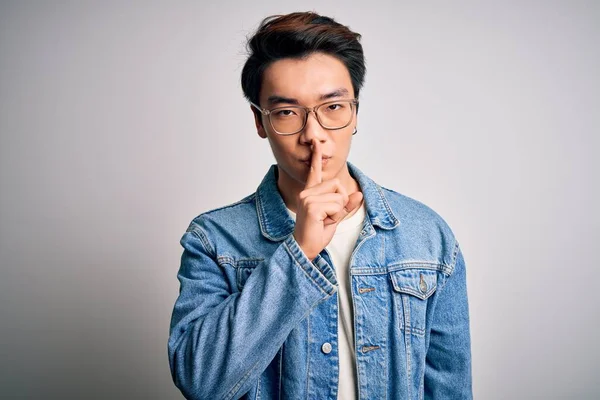 Young handsome chinese man wearing denim jacket and glasses over white background asking to be quiet with finger on lips. Silence and secret concept.