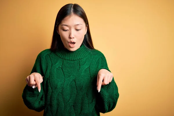 若いです美しいですアジアの女性身に着けています緑の冬のセーター上の黄色隔離された背景ポインティング指で示す広告 驚きの顔とオープン口 — ストック写真