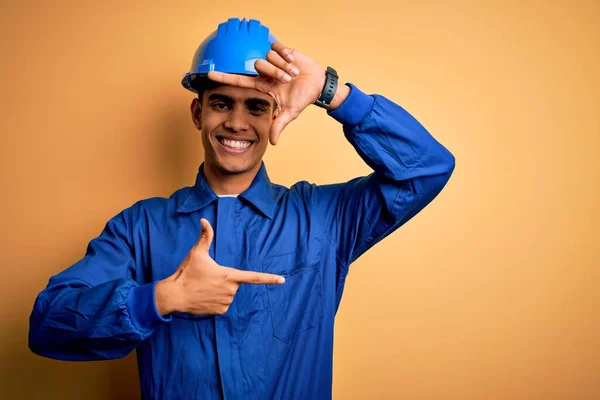 青い制服とセキュリティヘルメットを身に着けている若いハンサムなアフリカ系アメリカ人労働者の男性は幸せな顔で手と指でフレームを作り笑顔 創造性と写真の概念 — ストック写真