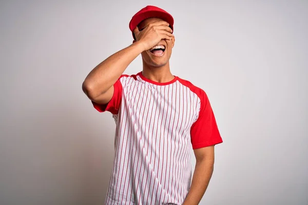 縞模様の野球Tシャツを着た若いハンサムなアフリカ系アメリカ人のスポーツマンが笑顔でキャップし 驚きのために目をカバー顔に手で笑っています ブラインドコンセプト — ストック写真