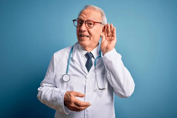 高个子灰发医生 头戴听诊器 身穿蓝色背景的医疗外套 面带微笑 两手相对地听着谣言或流言蜚语 失聪的概念 — 图库照片