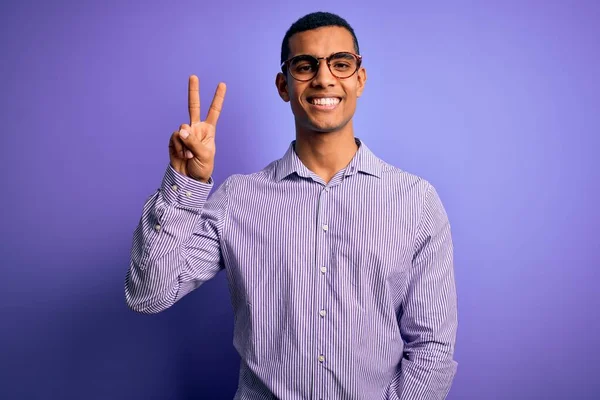 一位英俊的非洲裔美国男人 身穿条纹衬衫 戴着眼镜 背景是紫色的 手指头指向二号 面带微笑 自信而快乐 — 图库照片