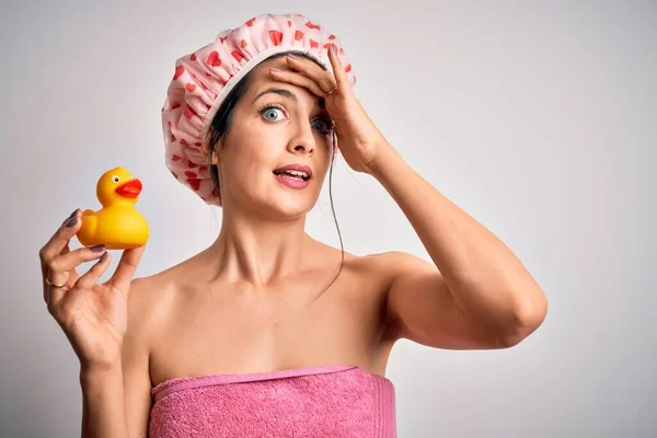 Young brunette woman with blue eyes wearing bath towel and shower cap holding duck toy stressed with hand on head, shocked with shame and surprise face, angry and frustrated. Fear and upset for mistake.