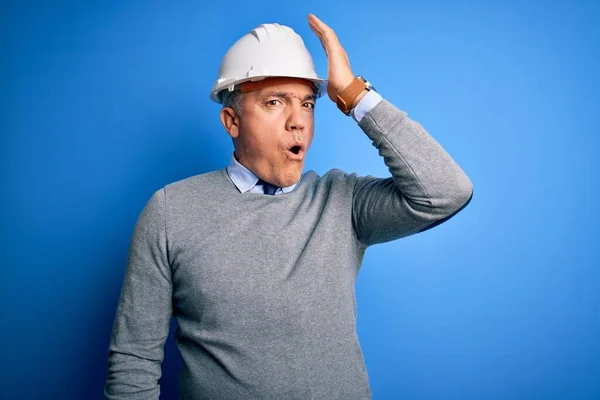Middle age handsome grey-haired engineer man wearing safety helmet over blue background surprised with hand on head for mistake, remember error. Forgot, bad memory concept.