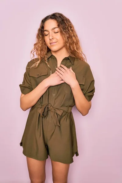 Young beautiful woman with blue eyes wearing casual summer dress over pink background smiling with hands on chest with closed eyes and grateful gesture on face. Health concept.