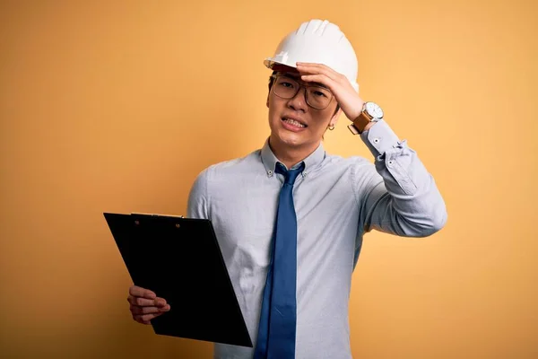 Young handsome chinese engineer man wearing safety helmet holding clipboard stressed with hand on head, shocked with shame and surprise face, angry and frustrated. Fear and upset for mistake.