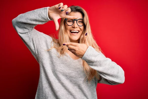 セーターと眼鏡を身に着けている若い美しいブロンドの女性は 幸せな顔をした手と指でフレームを作る孤立した赤い背景笑顔 創造性と写真の概念 — ストック写真