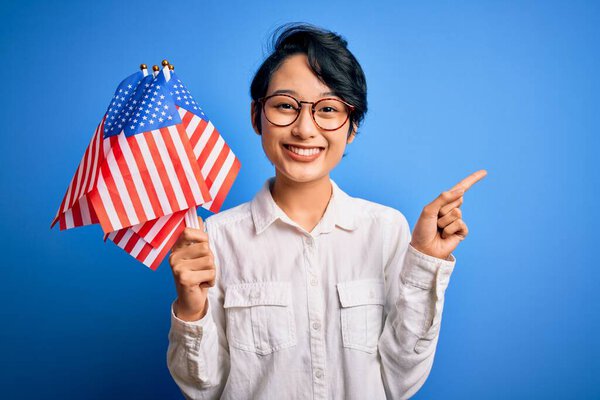 Young beautiful asian patriotic girl holding united states flags celebrating independence day surprised with an idea or question pointing finger with happy face, number one