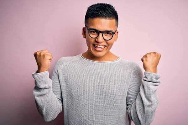 ピンクの背景にカジュアルなセーターと眼鏡を身に着けている若いハンサムなラテン人は 腕を上げて開いて目で成功したことに驚いて驚いて驚いています 勝者のコンセプト — ストック写真