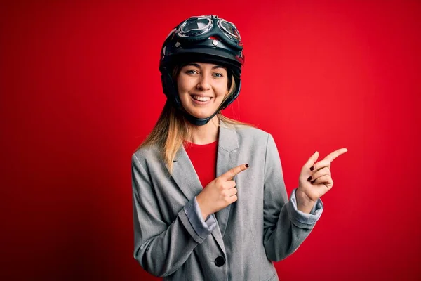 若いです美しいですブロンドオートバイの女性身に着けていますオートバイヘルメット以上赤背景笑顔と見ますカメラポインティングで二つの手と指で側面 — ストック写真