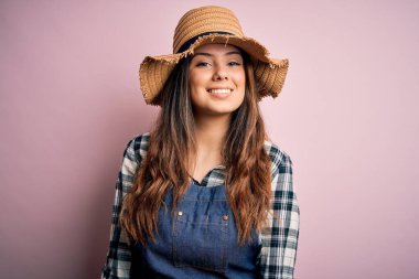 Genç, güzel esmer çiftçi kadın pembe arka planda önlük ve şapka takıyor ve yüzünde mutlu ve havalı bir gülümseme var. Şanslı kişi..