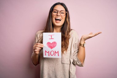 Elinde aşk dolu bir kağıt tutan genç bir kadın anneler gününü kutluyor çok mutlu ve heyecanlı, kazanan ifade zaferi kutluyor büyük bir gülümsemeyle bağırıyor ve ellerini kaldırıyor.