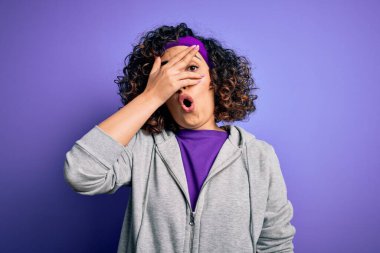 Güzel kıvırcık Arap sporcu kadın spor yapıyor. Mor arka planda spor giysisi giyiyor. Şok olmuş yüz ve gözleri, utanmış ifadeyle parmaklarının arasından bakıyor..