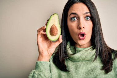Mavi gözlü, orta sağlıklı avokadoyu soyutlanmış beyaz arka planda tutan genç bir kadın. Şaşırmış bir yüzle korkmuş ve korku ifadesinden heyecanlanmış.