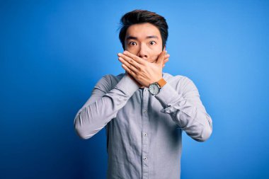 Genç, yakışıklı, sıradan bir tişört giyen, mavi arka planda duran Çinli bir adam yanlışlıkla ağzını kapatarak şok geçirdi. Gizli konsept.