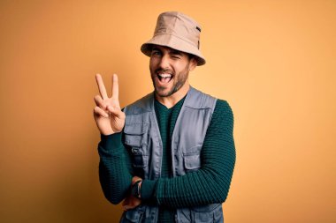 Tatilde sakallı yakışıklı turist sarı arka planda kaşif şapkası takıyor ve kameraya gülümsüyor parmaklarıyla zafer işareti yapıyor. İki numara..