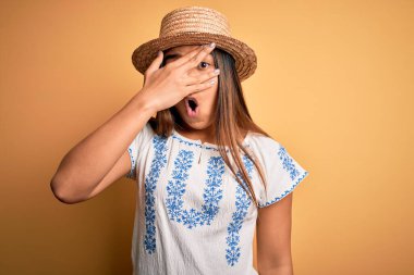 Genç ve güzel Asyalı bir kız günlük tişört ve şapka giyerek sarı arka planda duruyor. Şok olmuş bir şekilde yüzünü ve gözlerini kapatıyor. Utanmış bir ifadeyle parmaklarının arasından bakıyor..