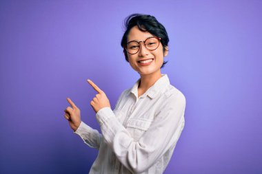 Genç, güzel Asyalı bir kız, günlük gömlek ve gözlük takıyor mor arka planda duruyor gülümsüyor ve kameraya bakıyor..