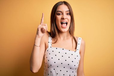 Sarı arka planda dikilmiş sıradan elbiseler giyen genç, güzel esmer kadın başarılı bir fikirle parmağını kaldırdı. Heyecanlı ve mutlu. Bir numara..