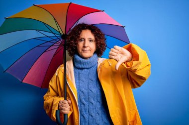 Orta yaşlı kıvırcık saçlı, yağmurluk giyen, mavi arka planda renkli şemsiyeli, kızgın yüzlü, olumsuz işaretli baş parmaklı, reddedilme kavramından hoşlanmayan bir kadın.