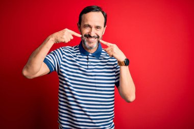 Orta yaşlı, beyaz, sıradan çizgili polo giyen, izole edilmiş kırmızı arka planda duran neşeli gülümseyerek parmaklarını ve ağzını gösteren bir adam. Diş sağlığı konsepti.