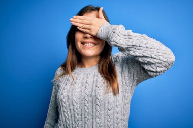 Güzel, genç bir kadın, mavi izole edilmiş arka planda dikilen yün bir süveter giyiyor. Gülümsüyor ve elleri yüzünde şaşkınlıktan gözleri kapatıyordu. Kör kavram.