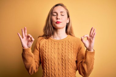Sarı arka planda sıradan bir süveter giyen genç ve güzel sarışın kadın rahatlamış ve parmaklarıyla meditasyon hareketi yaparken gözleri kapalı gülümsüyor. Yoga konsepti.