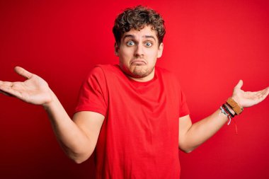 Sarışın, kıvırcık saçlı, kırmızı arka planda sıradan tişörtler giyen, elleri ve kolları havada, kafası karışmış genç, yakışıklı bir adam. Şüphe kavramı.