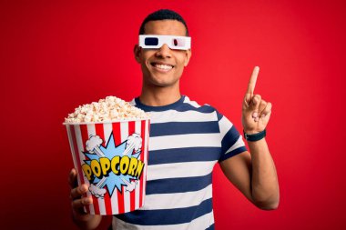 Genç ve yakışıklı Afro-Amerikalı adam film izlerken 3D gözlük takıyor yüzünde kocaman bir gülümsemeyle patlamış mısır yiyor ve kameraya el ile işaret ediyor..