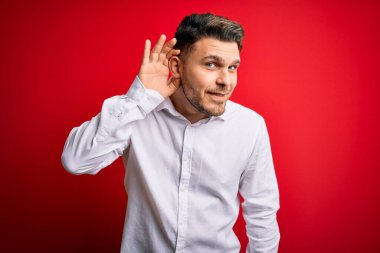 Mavi gözlü, zarif gömlekli genç bir iş adamı kırmızı izole edilmiş arka planda dikiliyor. Kulağından kulağa gülümsüyor. Dedikoduları ve dedikoduları dinliyor. Sağırlık kavramı.