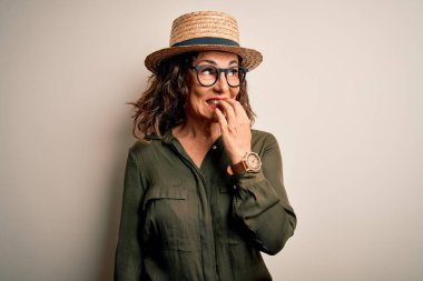 Orta yaşlı esmer kadın, gözlüklü ve şapkalı. İzole edilmiş beyaz arka planda duruyor. Elleri ağızdan tırnak yerken gergin ve stresli görünüyor. Endişe sorunu.