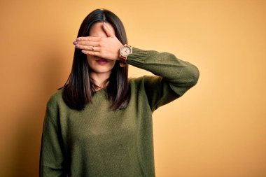 Mavi gözlü, sarı arka planda yeşil süveter giyen, gözleri elle kaplayan, ciddi ve üzgün görünen esmer bir kadın. Görme, saklama ve reddetme konsepti