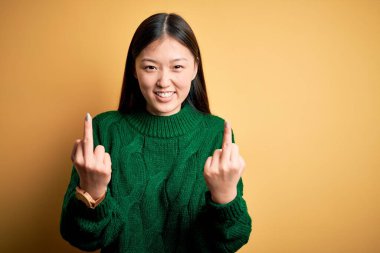 Young beautiful asian woman wearing green winter sweater over yellow isolated background Showing middle finger doing fuck you bad expression, provocation and rude attitude. Screaming excited