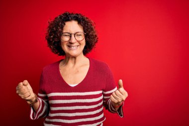 Orta yaşlı güzel kıvırcık saçlı kadın kırmızı arka planda sıradan çizgili süveter giyiyor. Çok mutlu ve heyecanlı. Kolları havada, gülümseyerek ve başarı için çığlık atarak kazanan jestler yapıyor. Kutlama konsepti.
