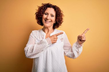 Orta yaşlı güzel kıvırcık saçlı kadın sarı arka planda rahat bir yaz elbisesi giyiyor gülümsüyor ve kameraya bakıyor iki eliyle ve parmaklarıyla yan tarafa bakıyor..