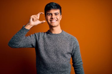İzole edilmiş turuncu arka plan üzerinde duran sıradan bir süveter giyen genç yakışıklı gülümseyen ve kendine güvenen el hareketleriyle küçük boy işareti yapan parmaklarıyla ve kamerasıyla. Ölçü kavramı.