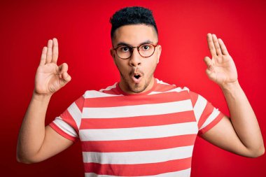 Sıradan çizgili tişört ve gözlük takan genç ve yakışıklı bir adam izole edilmiş kırmızı arka planda şaşırmış ve şaşırmış görünüyor. Tamam sembolünü parmaklarıyla yapıyor. Çılgın ifade