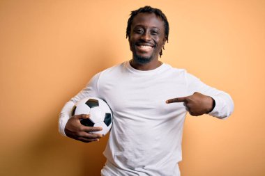 Genç Afrikalı Amerikalı oyuncu futbol topunu sarı arka planda tutarak oynuyor. Şaşırtıcı yüz ifadesiyle kendini işaret ediyor.