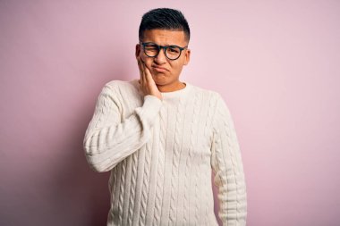 Beyaz kazak giyen ve pembe arka planda gözlük takan genç, yakışıklı Latin bir adam diş ağrısı ya da diş ağrısı yüzünden acı dolu bir ifadeyle dudaklarına dokunuyor. Dişçi