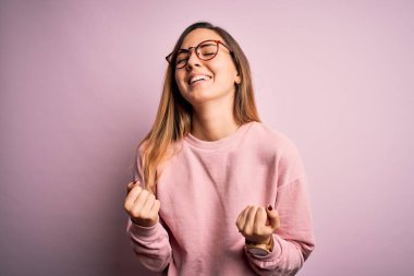 Mavi gözlü, pembe arka planda süveter ve gözlük takan güzel sarışın kadın çok mutlu ve heyecanlı bir şekilde kollarını kaldırıp, gülümseyip başarı için çığlık atıyor. Kutlama konsepti.