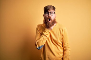 Sarı yalıtılmış arka planda gözlüklü, sakallı yakışıklı İrlandalı kızıl adam. İnançsızlığa, şaşkınlığa ve elleri çenesinde olan şaşkınlık ifadesine hayran görünüyor.