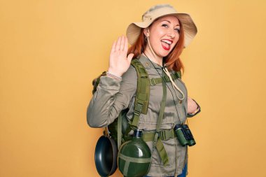 Sırt çantası ve sarı arka planda şapka takan genç kızıl saçlı kadın merhaba diyor mutlu ve güler yüzlü, dostça bir hoş geldin jesti